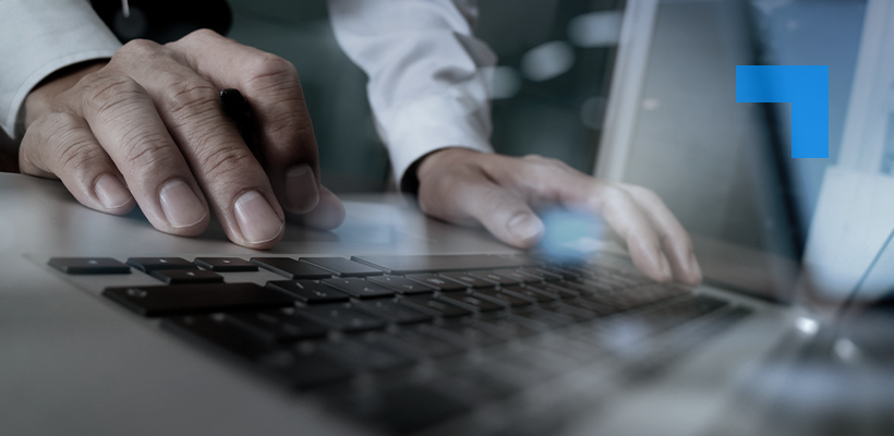 Close-up of a person using a laptop, with one hand on the keyboard and the other on a mouse. The individual is wearing a white shirt with a black sleeve, suggesting a professional setting.