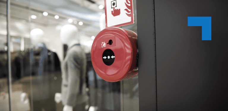 A red fire alarm activation button mounted on a wall in a modern retail store, with mannequins in suits visible in the background.