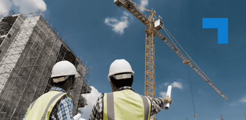 Two construction workers wearing hard hats and high-visibility vests look up at a tall yellow tower crane beside a large building covered in scaffolding. One worker points toward the crane while holding rolled-up plans.