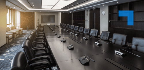 A large, modern conference room with a long, polished wood table surrounded by many black leather chairs. Individual microphones are placed at each seat. The room features recessed ceiling lighting, a wall-mounted flat-screen display at the far end, built-in wooden shelving, double wooden doors, and large windows on the left letting in warm sunlight.