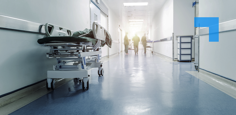 A hospital corridor with bright lighting, a vacant patient bed on the left, medical equipment along the walls, and blurred people walking towards the far end of the corridor.