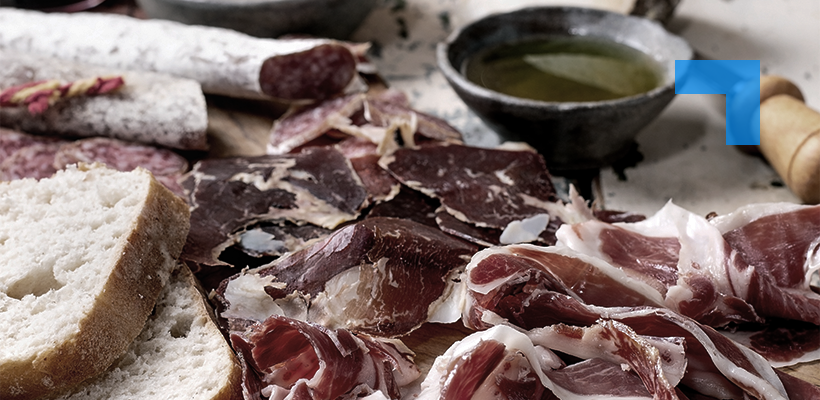 Assorted cured meats sliced and arranged on a wooden surface alongside crusty bread, salami, and a small bowl of olive oil, presented as a rustic charcuterie spread.