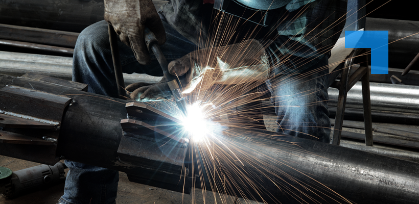 A worker wearing gloves welds a large metal pipe in an industrial workshop, with bright sparks flying from the welding torch.