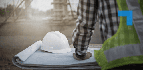 Construction professional wearing a high-visibility vest reviews blueprints on a site table, with a white hard hat resting on the plans and an industrial construction area in the background.