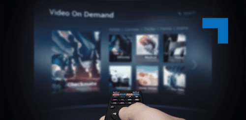 Hand holding a TV remote control pointed at a blurred video-on-demand streaming interface displaying movie thumbnails.