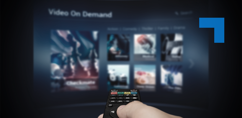 Hand holding a TV remote control pointed at a blurred video-on-demand streaming interface displaying movie thumbnails.