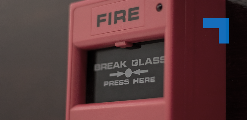 Close-up of a red wall-mounted fire alarm call point displaying the words “FIRE” and “BREAK GLASS – PRESS HERE.”