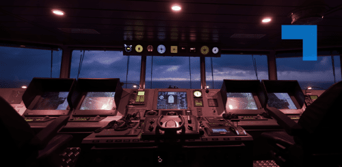 The interior of a ship’s bridge at dusk, showing multiple illuminated navigation screens, radar displays, control panels, and a central steering console, with the ocean and darkening sky visible through the front windows.