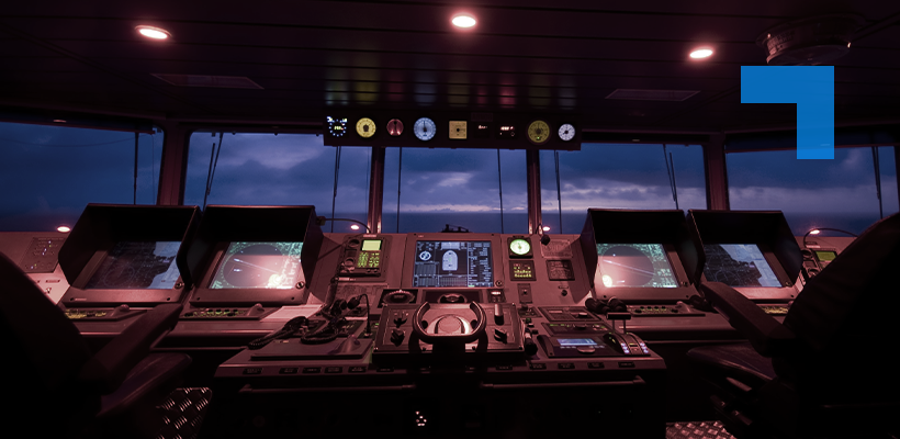 The interior of a ship’s bridge at dusk, showing multiple illuminated navigation screens, radar displays, control panels, and a central steering console, with the ocean and darkening sky visible through the front windows.