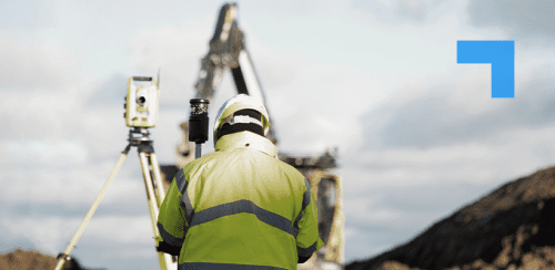 Surveyor in a high-visibility jacket and hard hat operating a tripod-mounted surveying instrument at a construction site, with an excavator and earthworks in the background under a cloudy sky.