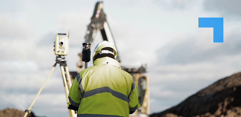 Surveyor in a high-visibility jacket and hard hat operating a tripod-mounted surveying instrument at a construction site, with an excavator and earthworks in the background under a cloudy sky.