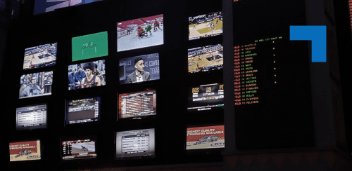 A wall of multiple TV screens in a dimly lit sportsbook or control room, each displaying different live sports events and broadcasts, alongside digital betting odds boards with scrolling numbers and team names.