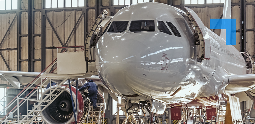 Front view of a commercial airplane inside a maintenance hangar, with its nose panels open and technicians working on equipment near the front landing gear and engine.