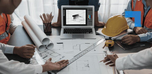 Architects and engineers reviewing building plans around a desk with a laptop displaying a 3D model, alongside blueprints, measuring tools and a hard hat, collaborating on a construction design project.