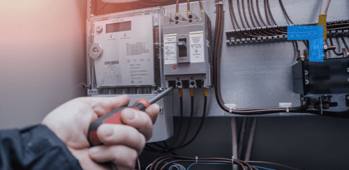 Close-up of a technician using a screwdriver to inspect or adjust an electrical meter and circuit breaker panel with visible wiring inside an industrial or commercial setting.
