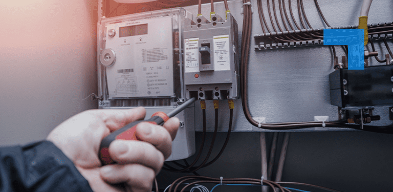 Close-up of a technician using a screwdriver to inspect or adjust an electrical meter and circuit breaker panel with visible wiring inside an industrial or commercial setting.