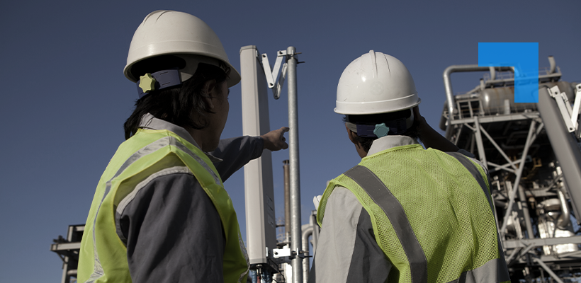 Two engineers in hard hats and high-visibility vests inspect industrial equipment at an outdoor facility, with one pointing toward a vertical structure.