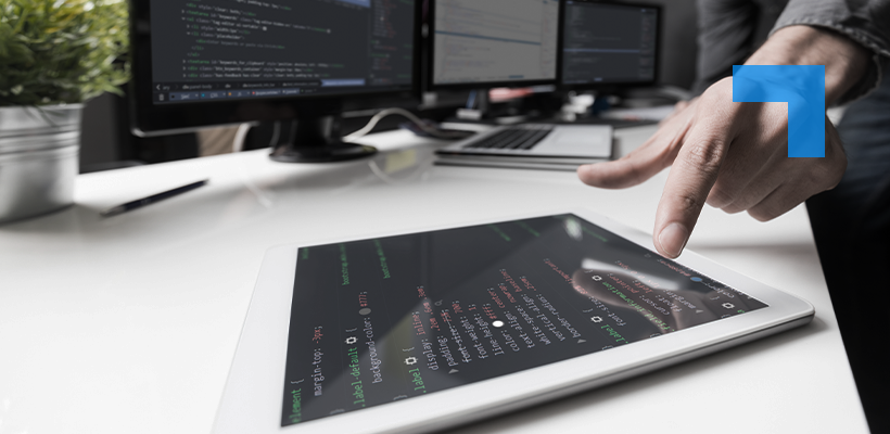 A person points at a tablet displaying lines of programming code on a desk beside multiple computer monitors showing software development screens in an office workspace.