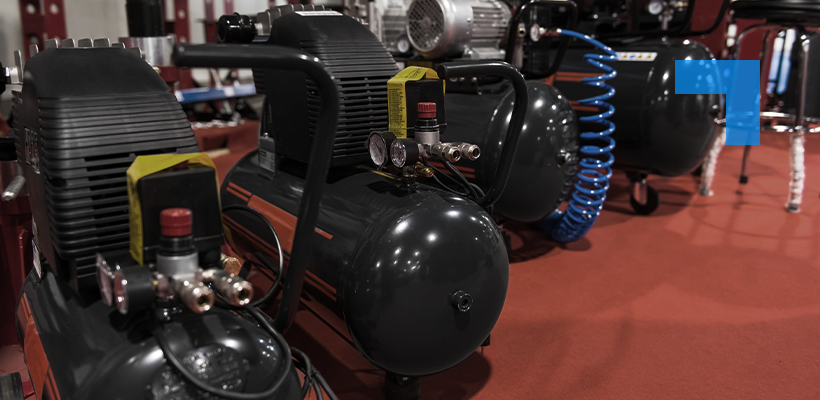 Industrial air compressors displayed on a showroom floor, with pressure gauges and coiled blue air hose visible in a warehouse setting.