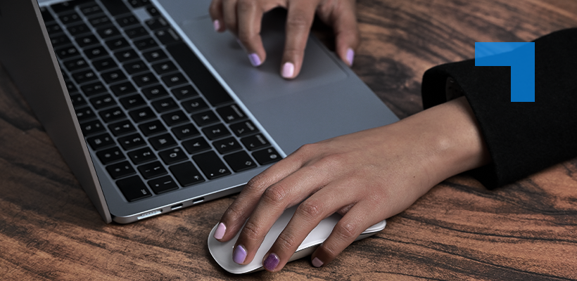 A person using a laptop and wireless mouse on a wooden desk, with one hand on the trackpad and the other on the mouse.