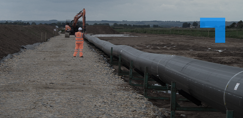 A worker in high-visibility PPE inspecting a large pipeline on supports at a construction site, with an excavator operating in the background.