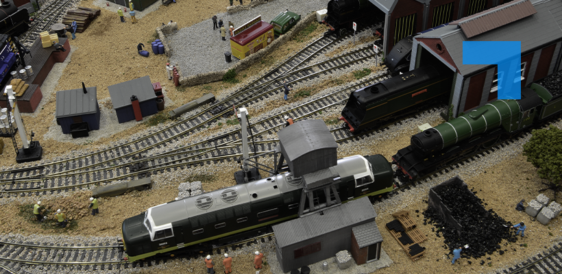 A detailed model railway layout featuring several vintage locomotives and freight cars around a busy rail yard, with multiple tracks, industrial buildings, workers, coal piles, and trackside equipment.