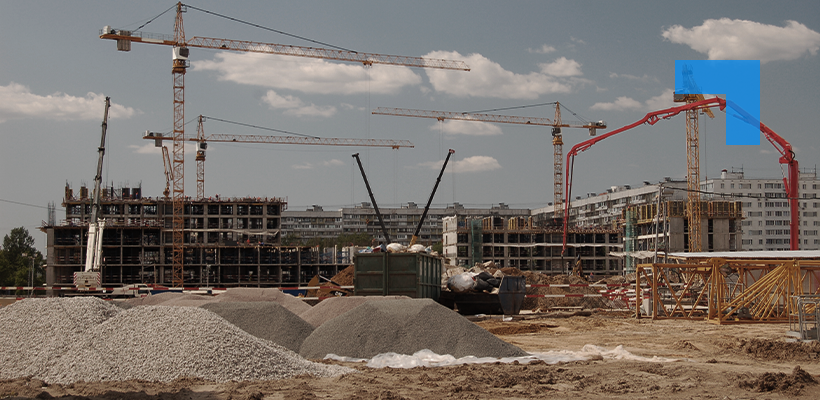 A large construction site with multiple tower cranes, partially built concrete structures, piles of gravel, and construction equipment under a partly cloudy sky.