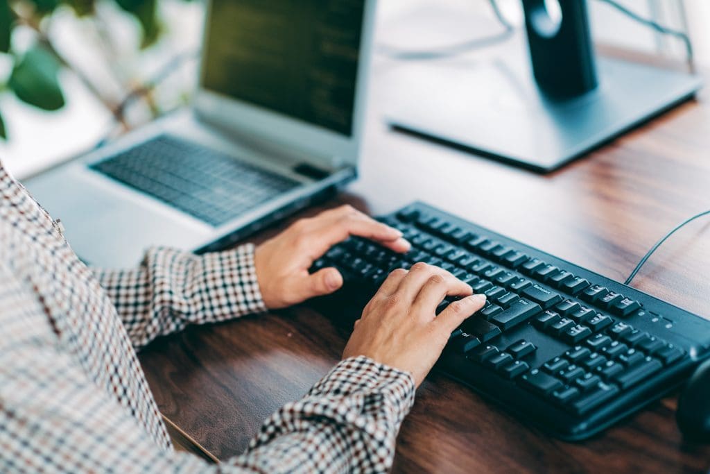 Close up of a man's hand typing on the a computer keyboard with a laptop at the side