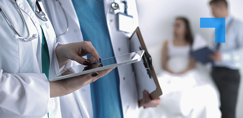 A clinical pharmacist in a white coat reviews patient information on a tablet, with a colleague consulting a patient in the background.