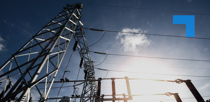A low-angle view looking up at a large steel electricity transmission pylon with high-voltage power lines stretching across a dramatic blue sky, with electrical substation equipment including insulators and switchgear visible in the foreground and sunlight breaking through the clouds.