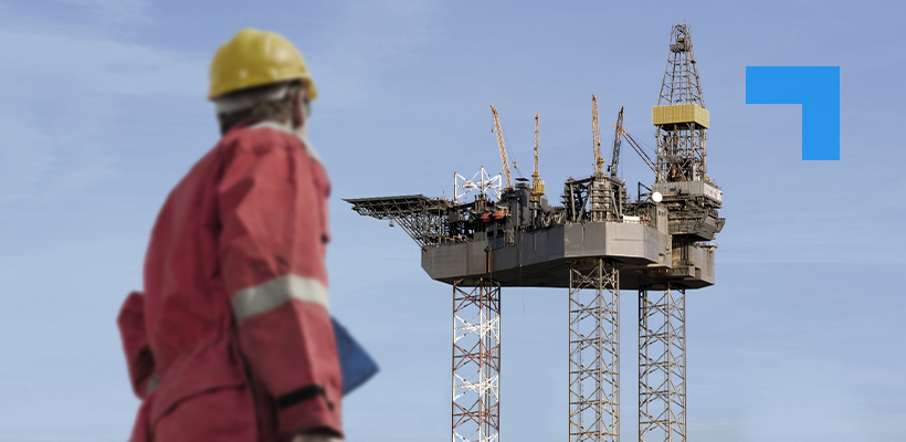 A worker in red high-visibility overalls and a yellow hard hat stands with their back to the camera, looking towards a large offshore oil and gas platform elevated on jack-up legs against a clear blue sky.